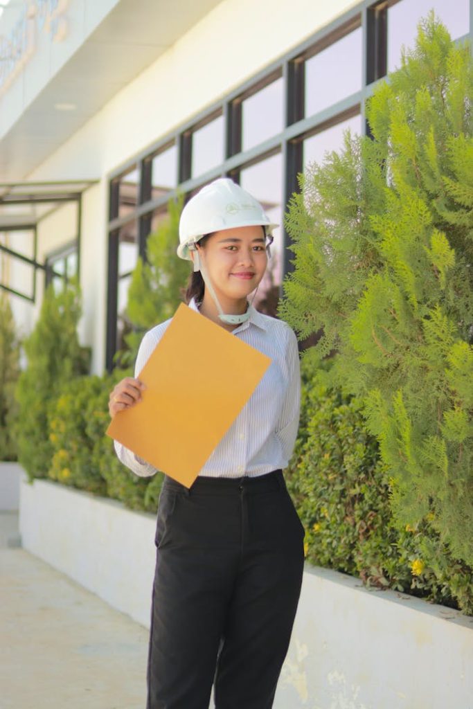 woman-in-white-shirt-and-black-pants-holding-yellow-paper-9399868 Asian female engineer wearing hard hat holding documents, smiling near greenery, standing outdoors.